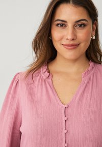 Pink textured blouse with a V-neck, soft ruffled collar, and five decorative buttons down the front. Loose-fitting sleeves.