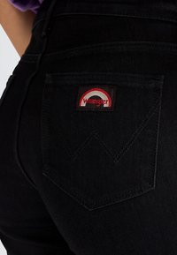 Nahaufnahme einer schwarzen Jeans-Tasche mit rotem und weißem Wrangler-Logo-Patch und eingenähter "W"-Detail.