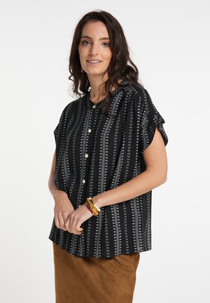 Frau mit dunklem lockigem Haar, die eine schwarze gemusterte kurzärmelige Bluse mit Knöpfen und einen braunen Rock trägt, lächelnd mit den Händen vor dem Körper verschränkt.
