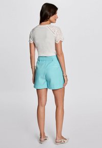Weißes Oberteil mit Spitze an den Ärmeln, kombiniert mit hellblauen Shorts. Die Shorts haben eine taillierte Passform und seitliche Taschen. Dazu werden schlichte Sandalen getragen.