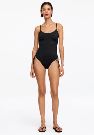 Femme debout portant un maillot de bain une pièce noir avec des liens latéraux réglables et des tongs noires sur un fond blanc uni.