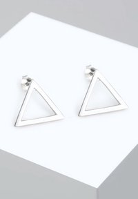 Paire de boucles d’oreilles clous en métal argenté en forme de triangle avec centre ouvert, sur une surface blanche et grise.