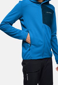 Felpa con zip blu realizzata in tessuto morbido, con un pannello centrale blu scuro, due tasche laterali e cappuccio regolabile. Caratterizzata dal logo VAUDE.