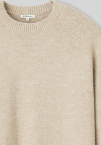 Pull beige oversize avec col rond côtelé, texture douce et détails minimalistes. Le tissu semble chaud et confortable. Étiquette de taille visible.