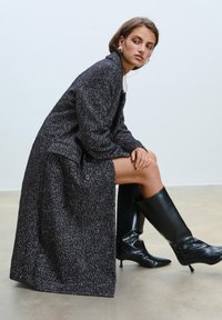 Cappotto lungo bianco e nero a spina di pesce con dettaglio di un solo bottone, indossato sopra un vestito lungo fino alla coscia, abbinato a stivali neri in pelle con tacco alto.