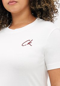 Άσπρη βαμβακερή μπλούζα με στρογγυλή λαιμόκοψη και κοντά μανίκια, με κεντημένο λογότυπο "C.K." σε μπορντό στο στήθος.