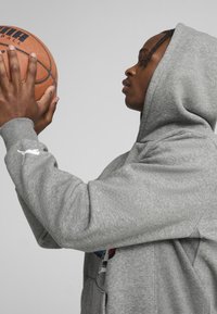 Grijze hoodie met een voorvak, textuurstof en een witte logo op de mouw. Iemand houdt een basketbal vast in zijaanzicht.