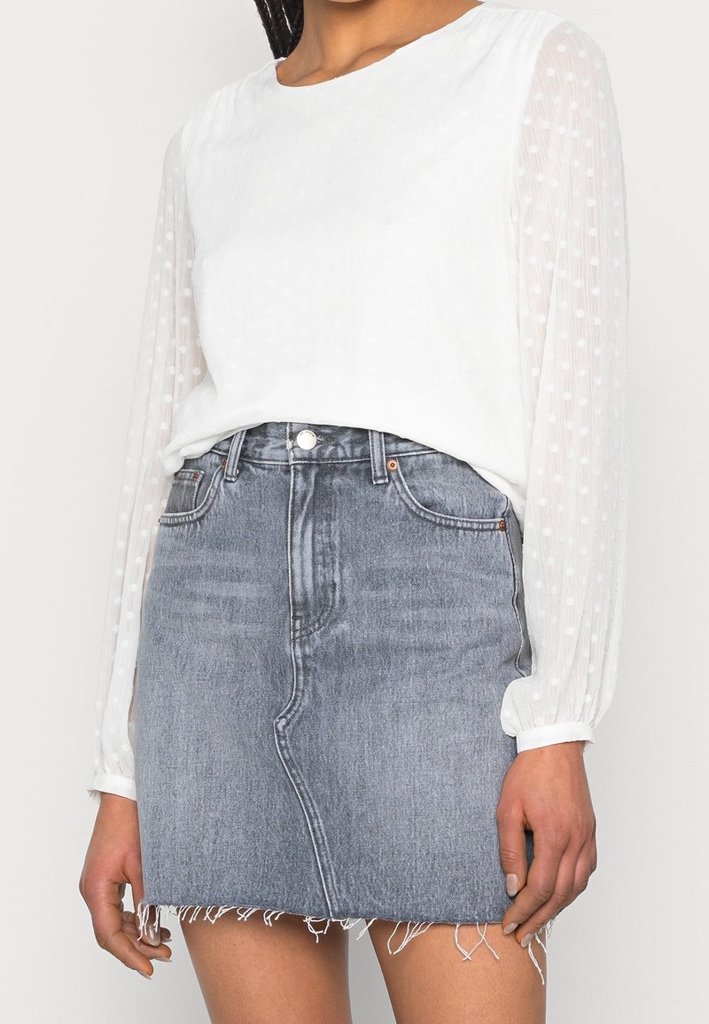 Femme portant un chemisier blanc à manches longues transparentes à pois et une jupe en jean bleu clair taille haute avec un ourlet effiloché.