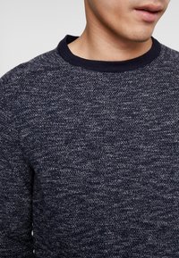 Man som bär en mörkblå och vit prickig stickad tröja med en enfärgad mörkblå ribbad rund hals.