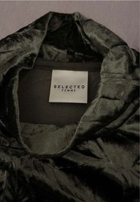 Dunkelgrüner, weicher Samtstoff mit einem tiefen Ausschnitt. Verfügt über ein weißes Etikett mit dem Text "SELECTED FEMME". Elegante Textur und Glanz.