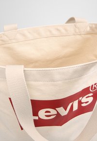 Torba na zakupy wykonana z płótna w kremowym kolorze, z czerwonym logo Levi's i teksturowaną tkaniną. Zawiera szerokie, płaskie paski na ramię do noszenia.