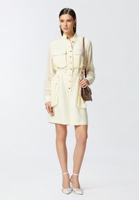 Abito button-up in tessuto giallo chiaro, con due tasche sul petto, vita elastica e tasche a scomparsa. Abbinato a sandali con tacco bianchi.