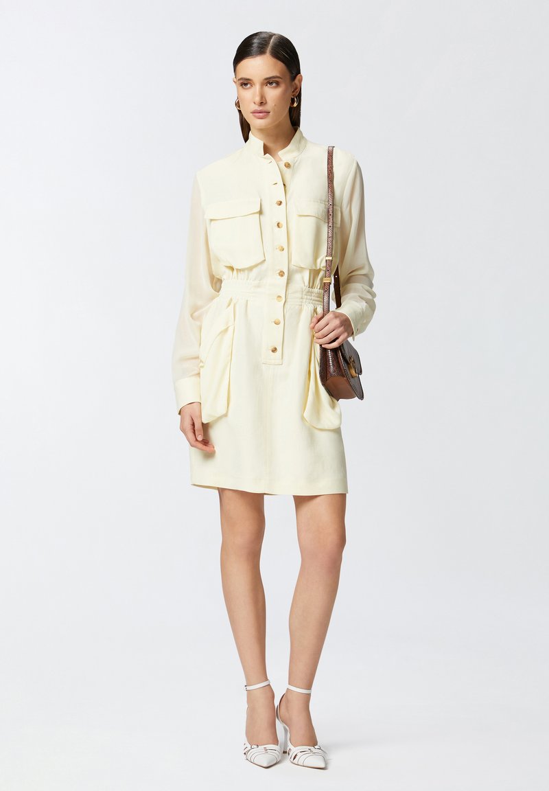 Abito button-up in tessuto giallo chiaro, con due tasche sul petto, vita elastica e tasche a scomparsa. Abbinato a sandali con tacco bianchi.