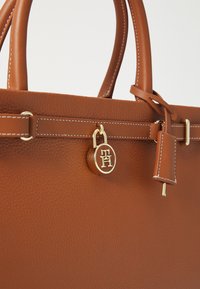 Ruda tekstūruota odinė nešiojamoji rankinė su auksinėmis detalėmis, turinti apvalų logotipą ir akcentines siuvimo detales ant rankenų bei kūno.