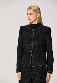 Veste en tweed noir au design structuré, avec un col rond, des accents en fil doré et cinq boutons sur le devant.