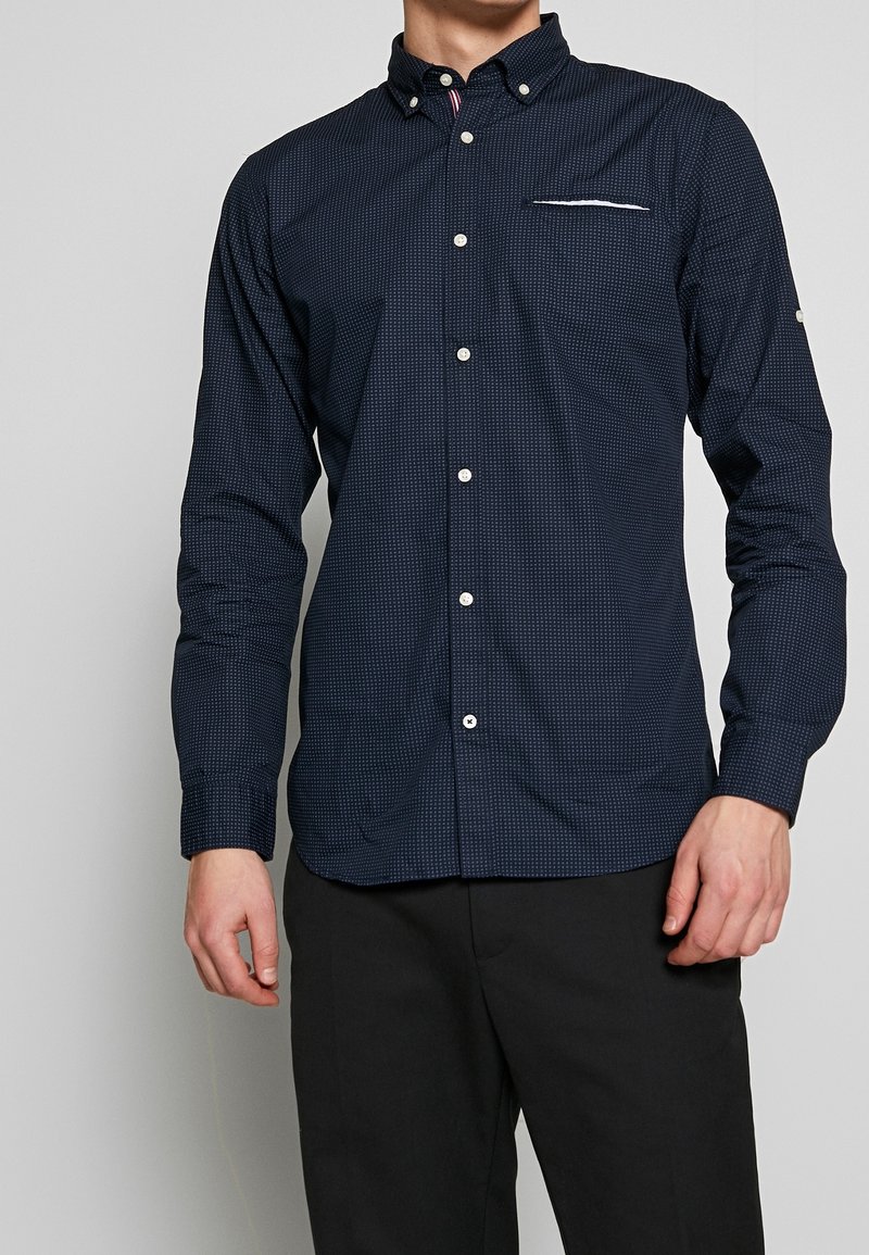 Homme portant une chemise bleu marine à boutons avec un petit motif carré, col boutonné, et un pantalon noir sur un fond gris uni.