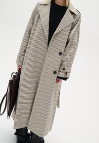 Cappotto lungo a quadri beige e blu navy con ampi rever, chiusura doppiopetto e spacchi laterali; abbinato a un maglione nero e a una borsa a spalla marrone.
