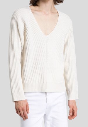 White ribbed V-neck sweater worn by a person with long sleeves, paired with white pants against a plain gray background.