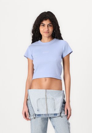 T-shirt court bleu clair avec une texture côtelée et des manches courtes, arborant un logo discret au centre. Porté avec des accents en denim.