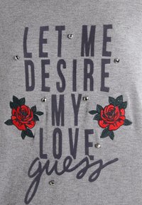 T-shirt gris affichant un texte sombre et audacieux "LAISSE-MOI DÉSIRER MON AMOUR" avec des roses rouges brodées et des ornements argentés répartis sur tout le design.