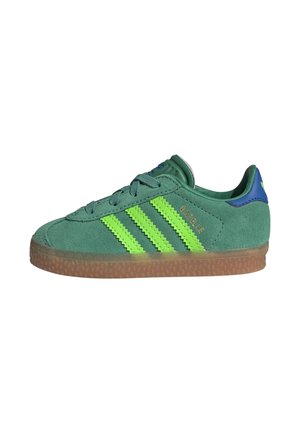 Zielony zamszowy sneaker z neonowo zielonymi paskami, brązową gumową podeszwą, niebieską zakładką na pięcie i napisem "GAZELLE" po boku.
