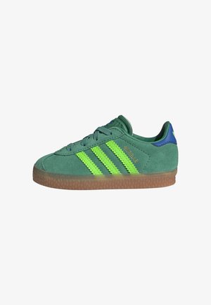 Zielony zamszowy sneaker z neonowo zielonymi paskami, brązową gumową podeszwą, niebieską zakładką na pięcie i napisem "GAZELLE" po boku.