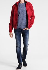 Rote leichte Jacke mit Reißverschluss über einem blauen Strickpullover, kombiniert mit blauen Jeans und dunklen Schuhen. Lässiges Design, taillierte Silhouette.
