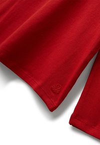 Červené okraje rukávů s neat švem a malým, nenápadným vyraženým logem blízko lemu na jednobarevném pozadí.