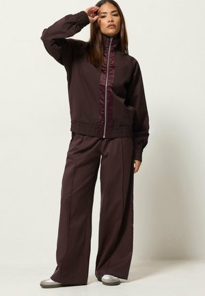 Femme debout portant une veste zippée marron foncé et un pantalon à jambes larges, chaussée de chaussures blanches et grises, avec une main levée sur le front.