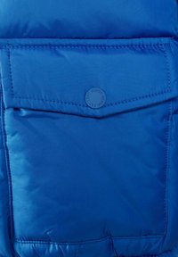 Mėlyna izoliuota striukė su priekiniais kišeniais ir apvaliu logotipu. Medžiaga yra lygi su minkšta, pūkuota tekstūra.