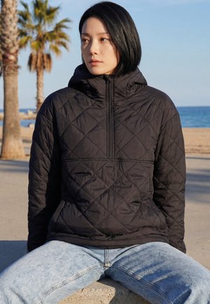 Fiatal nő fekete steppelt dzsekiben és világoskék farmernadrágban ül a szabadban, a háttérben tengerpart, pálmafák és az óceán látható.