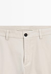 Lys beige bukser med en glat tekstur, forsynet med en sort knaplukning, bæltestropper og et rent, enkelt design.