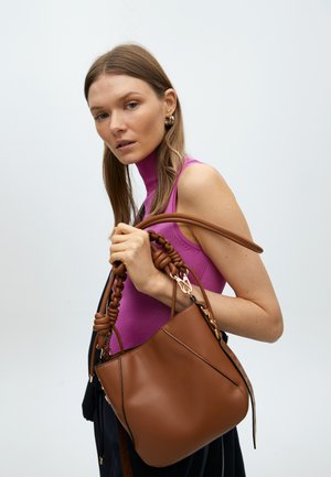 Borsa a mano in pelle marrone con manico intrecciato, texture liscia e dettagli in acciaio inox dorato, tenuta da una persona con una maglietta viola.