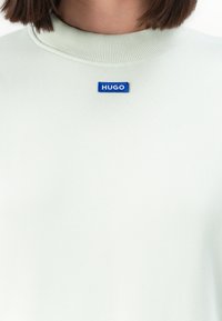 Lähikuva henkilöstä, joka pitää päällään vaaleanväristä ribbikaulusta, jossa on pieni sininen "HUGO"-merkki keskikauluksessa.