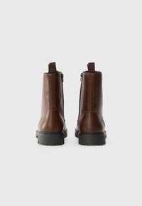 Botas de cuero marrón hasta el tobillo con una textura suave, forma recta, cremallera trasera y asas de bucle. Suelas de goma negra con detalle de banda de rodadura.