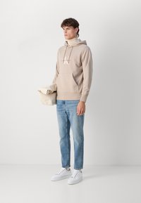 Felpa beige con tasca frontale e cordino, abbinata a jeans blu chiaro e sneaker bianche. Tessuto morbido e design minimalista.