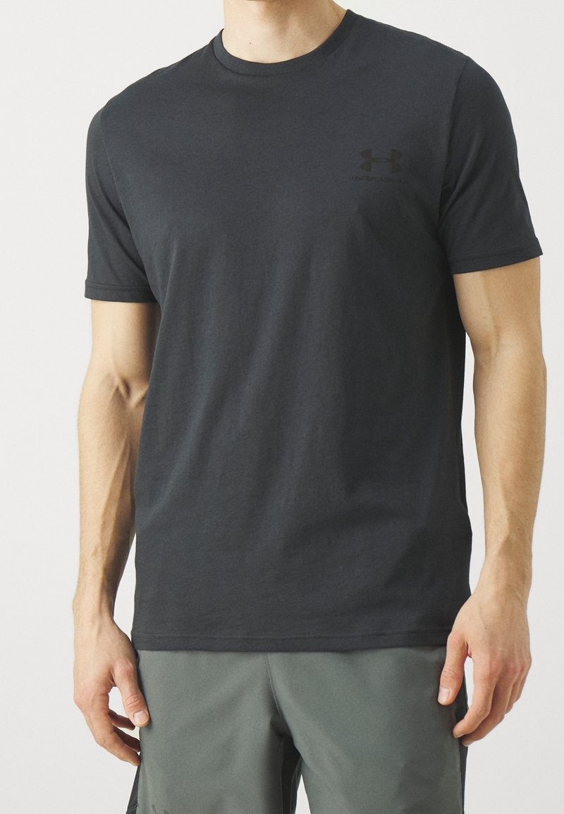Homme portant un t-shirt gris foncé uni à manches courtes avec un petit logo Under Armour sur la poitrine gauche et un short gris, debout, les bras détendus.