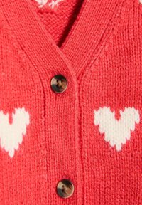 Czerwony sweter z dzianiny z białymi wzorami serc, dekoltem w kształcie litery V i brązowymi guzikami. Miękka struktura z widocznymi detalami szycia.
