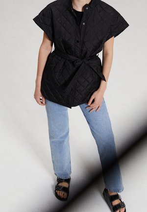 Person wearing a black quilted sleeveless jacket with belt, light blue jeans, and black sandals, standing against a plain light background.