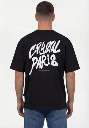 Mann trägt schwarzes T-Shirt mit großem weißen Schriftzug "CRYSTAL PARIS" auf dem Rücken, kombiniert mit blauen Jeans, mit dem Rücken zur Kamera.