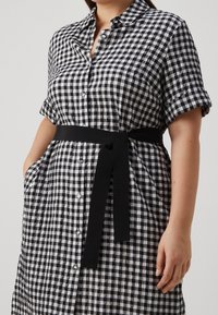 Černo-bíl dress s kostkovaným vzorem, límečkem, krátkými rukávy, knoflíky po celé přední části a černým textilním páskem. Obsahuje boční kapsy.