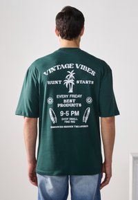 Dunkelgrünes, übergroßes T-Shirt mit weißem Druck, Palmbaumdesign und Text, der für einen Vintage-Markt wirbt, mit kurzen Ärmeln und lässigem Schnitt.