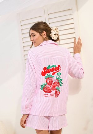Kvinde med brunt hår i en cremehårstrikker bærer lyserødt stribet pyjamas med en jordbærgrafik og teksten "Berry Sweet" på ryggen.