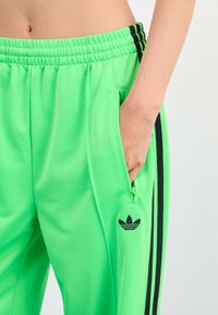 Green athletic pants made of smooth, lightweight fabric. Features black side stripes, zippered pocket, and a small black logo on the thigh.