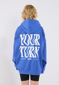 Felpa oversize blu con testo bianco "YOUR TURN" sulla schiena. Caratterizzata da un cappuccio con cordino e polsini a coste, realizzata in un tessuto morbido.