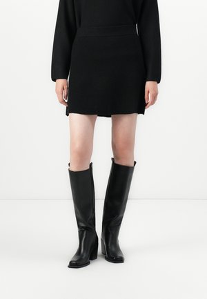 Γυναίκα που φοράει μαύρο φόρεμα με μακριά μανίκια και μαύρες δερμάτινες μπότες με τακούνι έως το γόνατο, ακουμπώντας σε απλό λευκό φόντο.