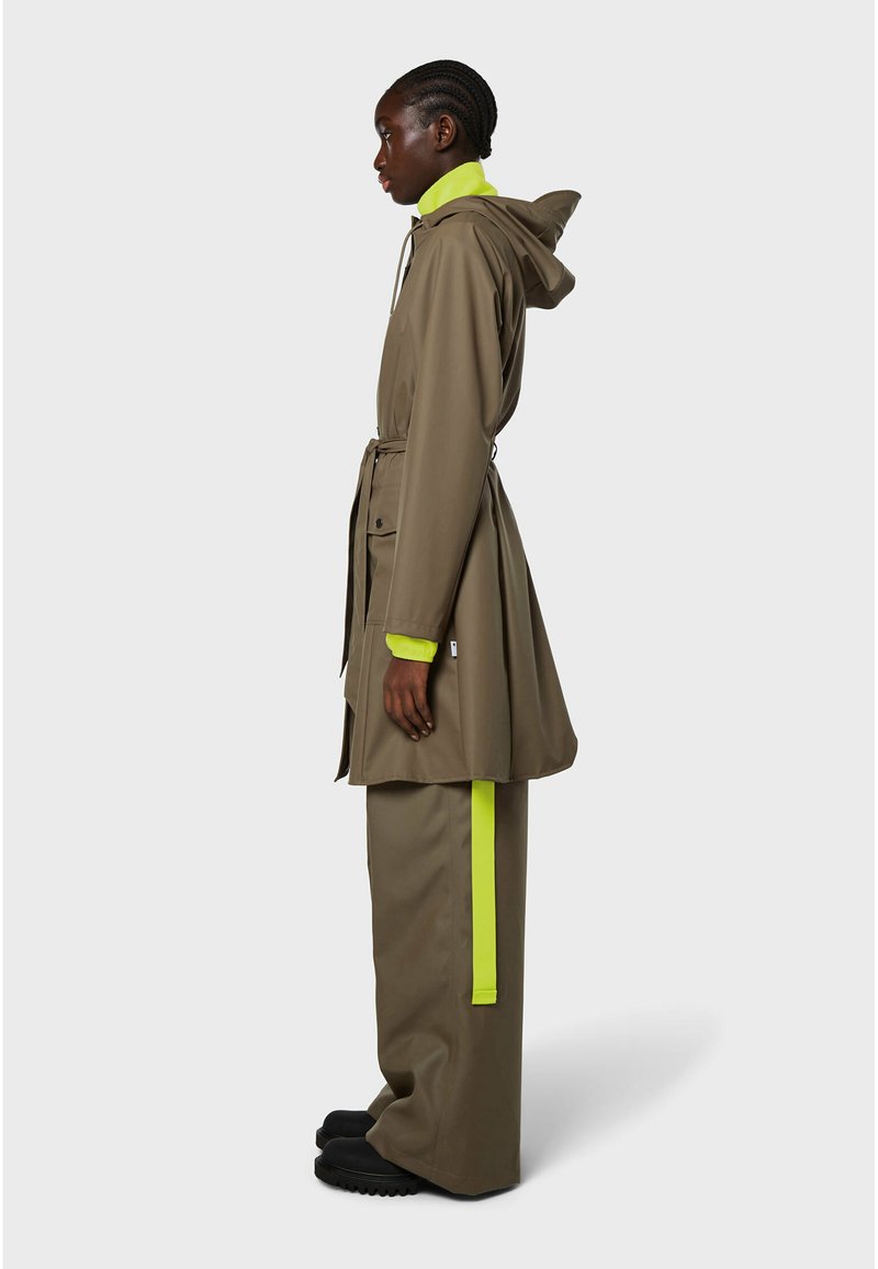 Impermeabile verde oliva con cappuccio, vita fascettata e dettagli verde neon. Abbinato a pantaloni larghi coordinati e calzature nere. Materiale liscio e impermeabile.
