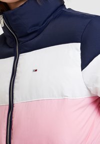 Color-blocked puffer jacket in navy, white, and pink. Features high collar, zip closure, and embroidered logo on the chest. Smooth fabric texture.