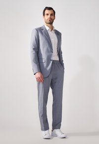 Hellgrauer Anzug mit kariertem Muster. Einreihiger Blazer, darunter ein weißes Hemd und elastische Hosen. Kombiniert mit weißen Sneakers.