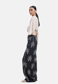 Licht beige cropped top met losse mouwen, gecombineerd met navy broek met witte palmboom borduring. Model staat in profiel.